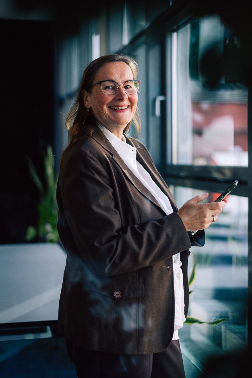 Beraterin mit Brille und Blazer, lächelnd mit Handy am Fenster bei grynia consulting