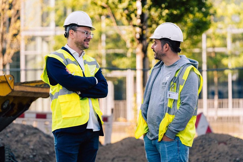 Zwei Mitarbeiter in Warnwesten und Helmen im Gespräch auf der Baustelle
